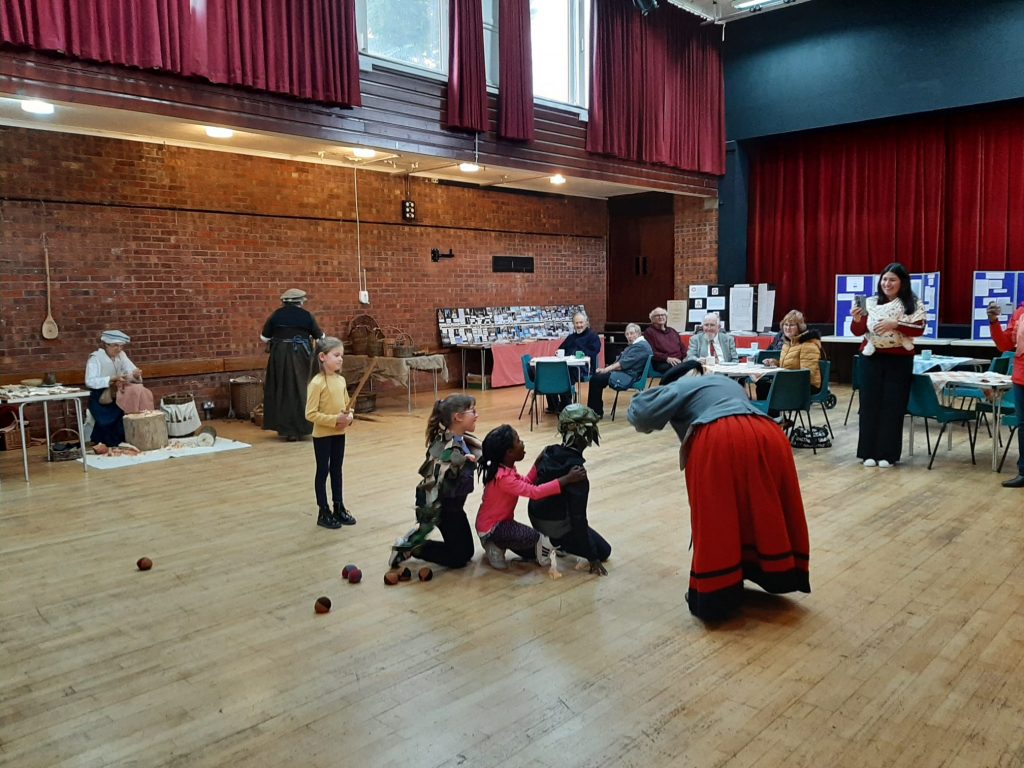 Emma bent over towards some children as they enact a play together with a dragon mask and sword