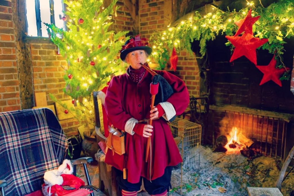 Emma playing the bagpipes dressed as a victorian standing in front of a christmas tree and an open fire. cosy and festive.