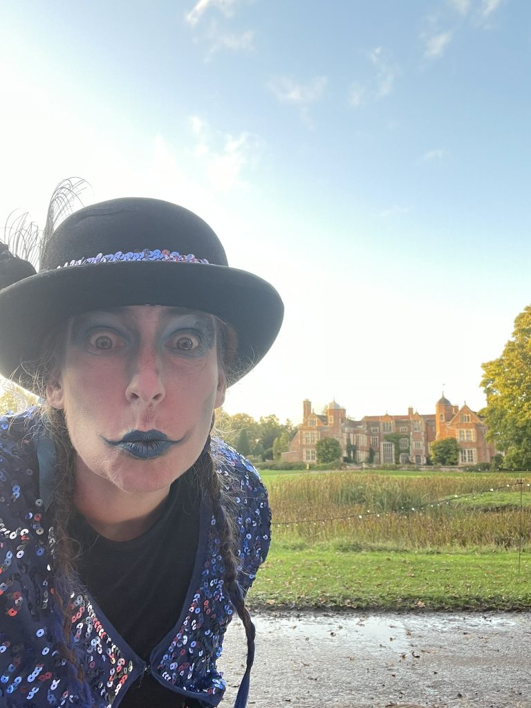 A picture of Emma wearing a black bowler hat and blue sequined outfit, with halloween makeup, in front of Kentwell Hall.