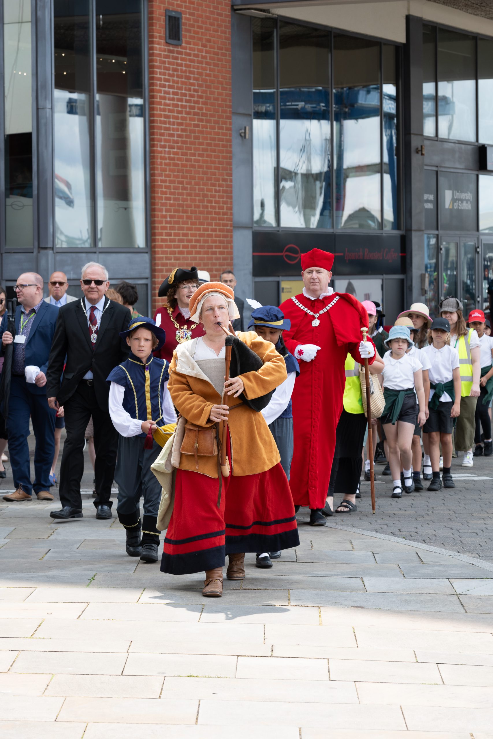 Emma dressed in tudor period clothing, playing the bagpipes, leads a parade of school children, along with Ipswich Mayor and a man dressed in red as Cardinal Wolsey.