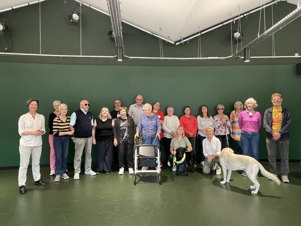 The Unscene singers stand in front of a green wall, 20 members strong, and two guide dogs in the front of the image. 
