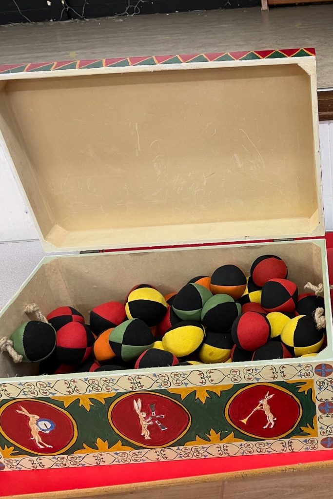 a wooden box painted in a medieval style, with an open lid, showing the contents of lots of juggling balls of various colours.  Next to this is a large willow basket.