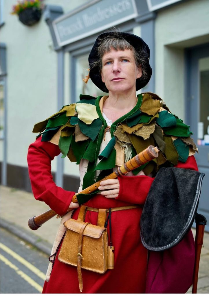 Emma stood holding a rauschpfeife, dressed in a red sudor dress with a green leafy cape and black hat